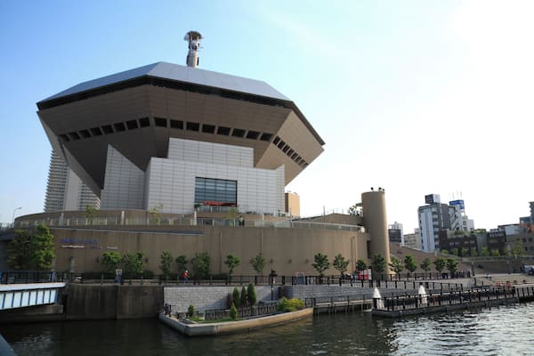 A modern building with a unique geometric design, situated by a waterfront with a bridge and trees.