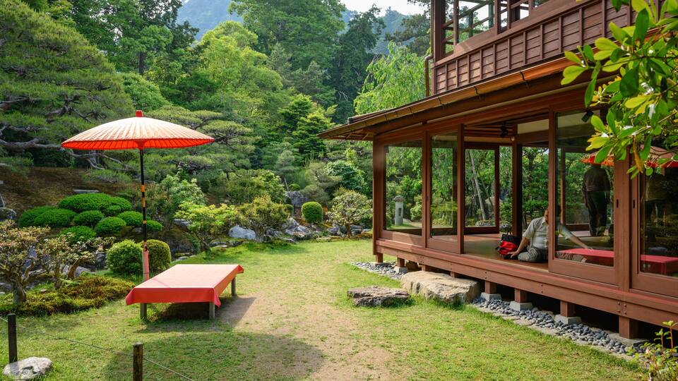 A traditional Japanese garden with a wooden house, a red umbrella, and a stone lantern.