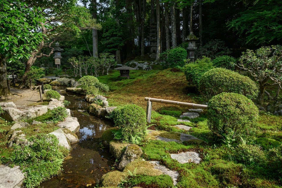 Un sereno jardí japonès amb un petit rierol, roques cobertes de molsa i fanals de pedra tradicionals.