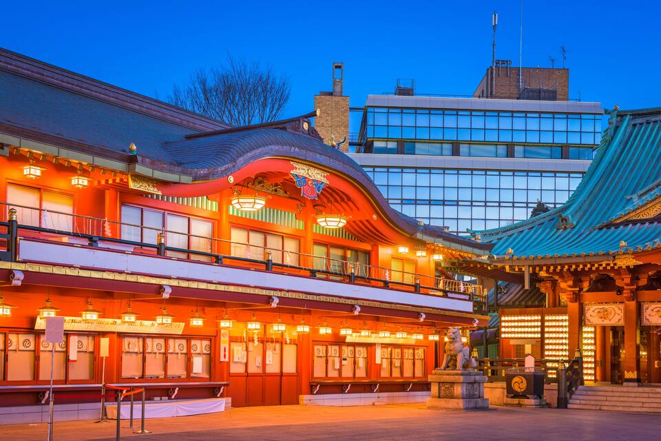 A traditional building with a curved roof and illuminated lanterns, adjacent to a modern glass building.