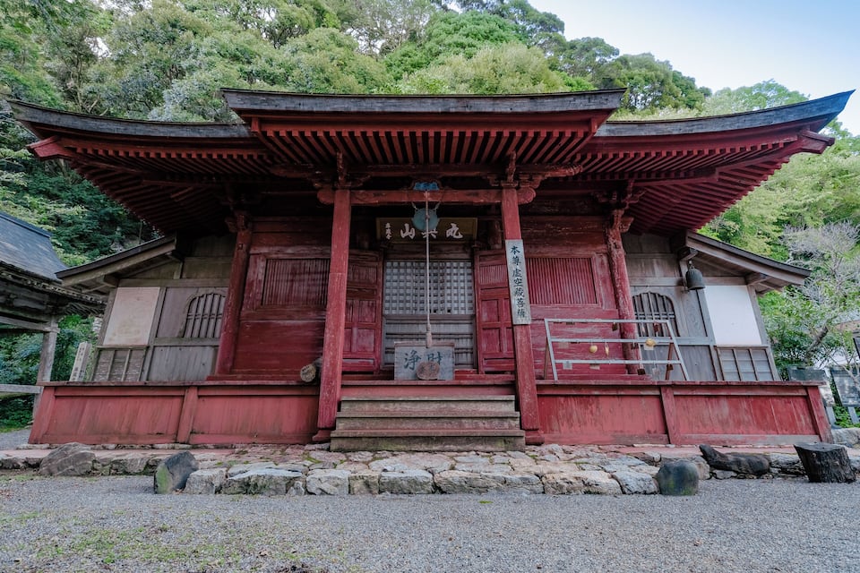 A traditional Japanese temple with red wooden architecture, a stone pathway, and a sign with Japanese characters.