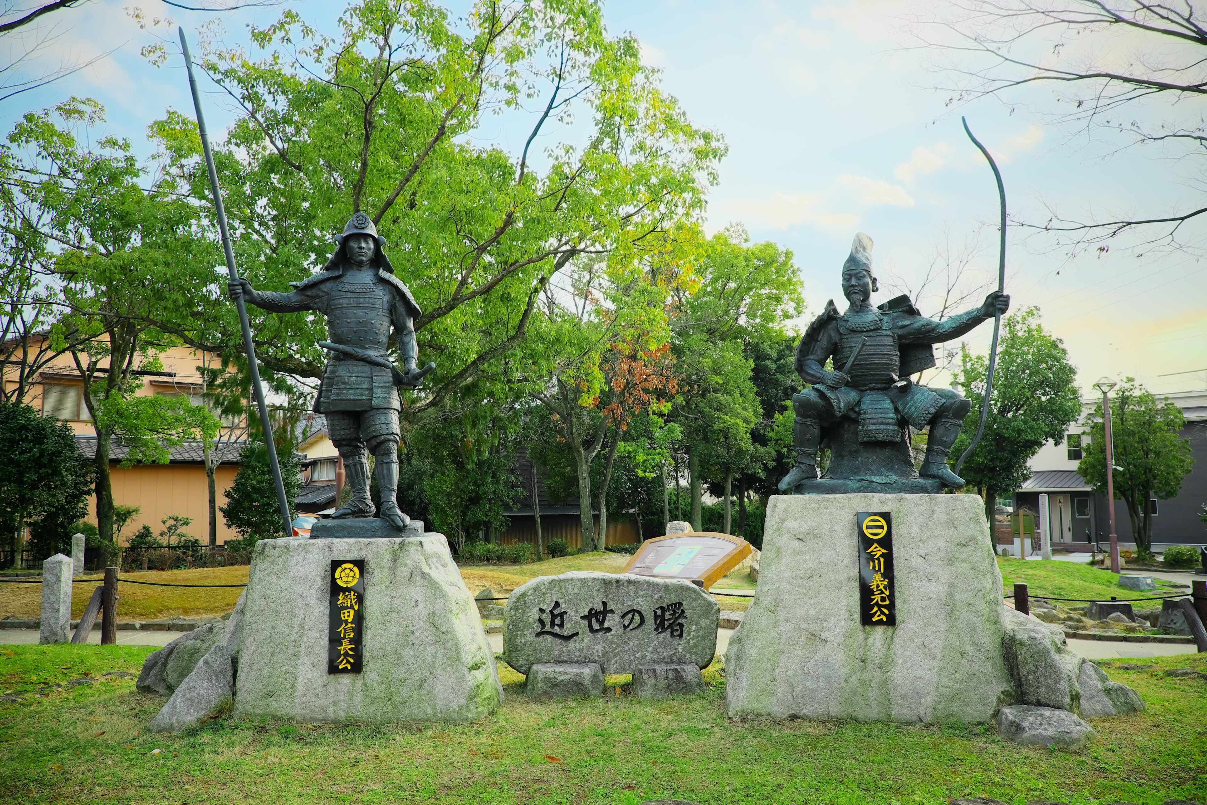 Two statues of samurai on stone bases with inscriptions, surrounded by trees and a stone monument in a park.