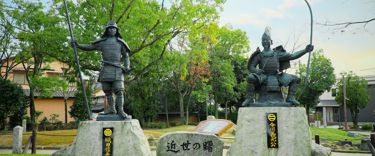 Two statues of samurai on stone bases with inscriptions, surrounded by trees and a stone monument in a park.