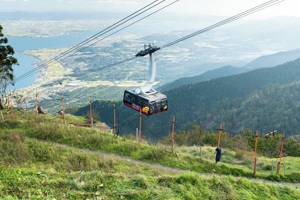 Un telefèric que avança sobre un paisatge montanyós amb vista a una vall i un cos d'aigua.