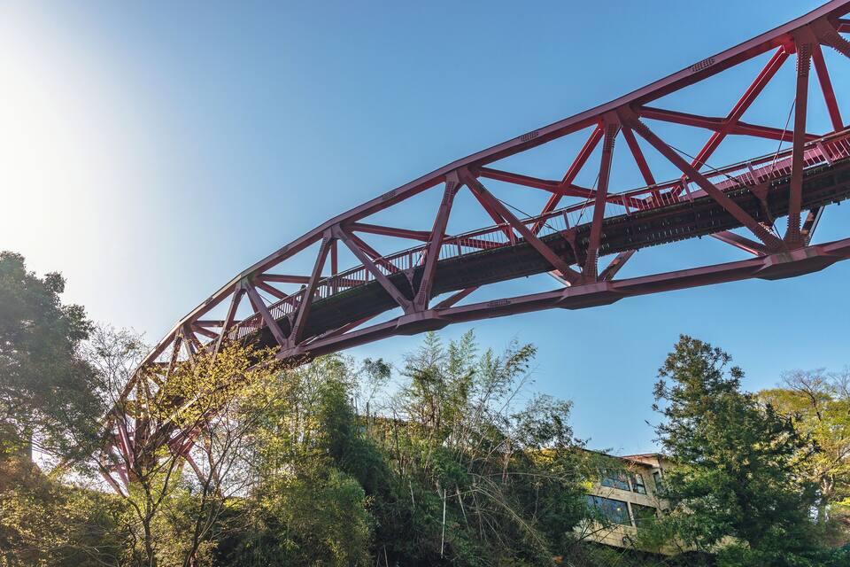 Uma ponte de aço vermelha com estrutura de treliça complexa, rodeada por vegetação verde densa e um edifício ao fundo.