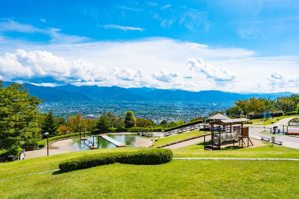Ein Park mit einem Spielplatz, einem kleinen Teich und Blick auf ferne Berge sowie eine Stadt.