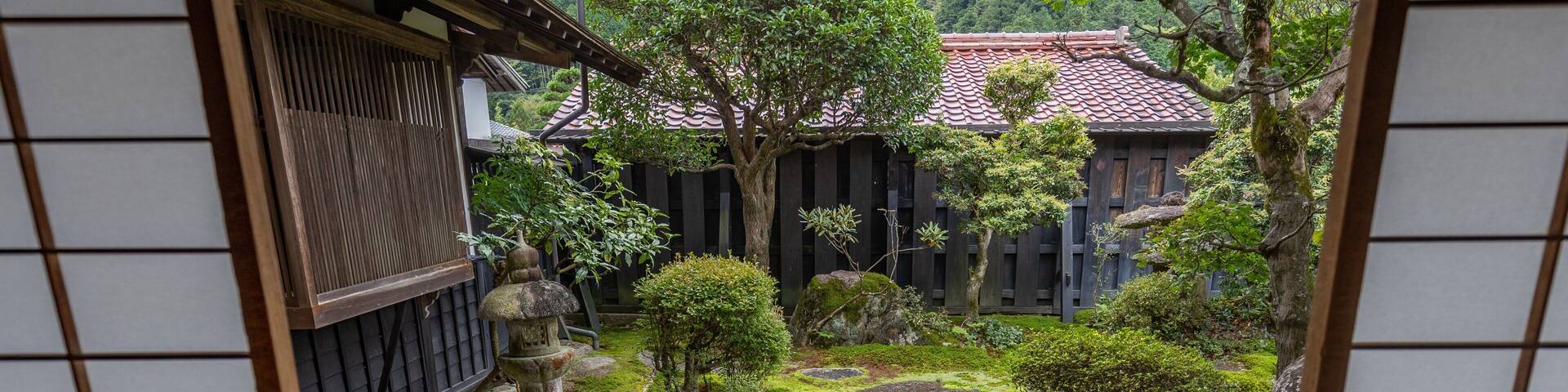 A traditional Japanese garden with moss-covered rocks, a stone lantern, and a wooden building with a tiled roof.