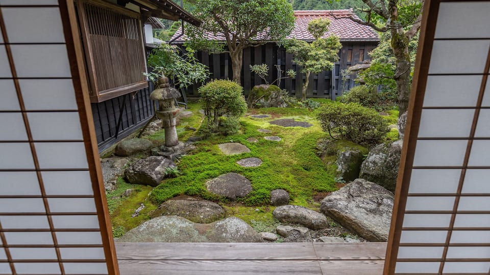 A traditional Japanese garden with moss-covered rocks, a stone lantern, and a wooden building with a tiled roof.