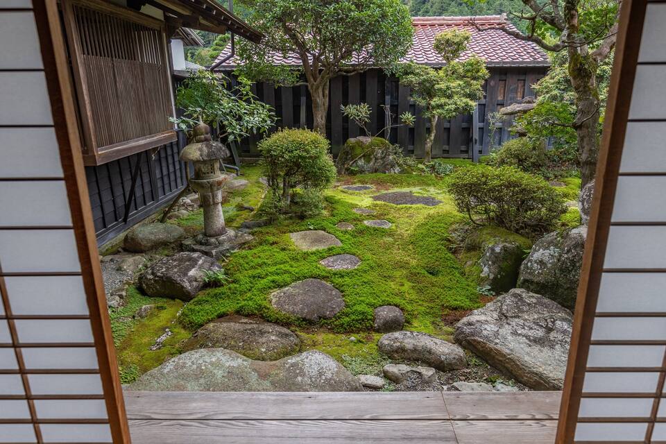 Um jardim japonês tradicional com pedras cobertas de musgo, um lança-pedras de pedra e um edifício de madeira com telhado em telhas.