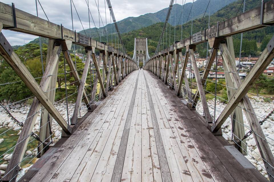 Un pont de fusta amb cables metàl·lics, envoltat per un riu i muntanyes.