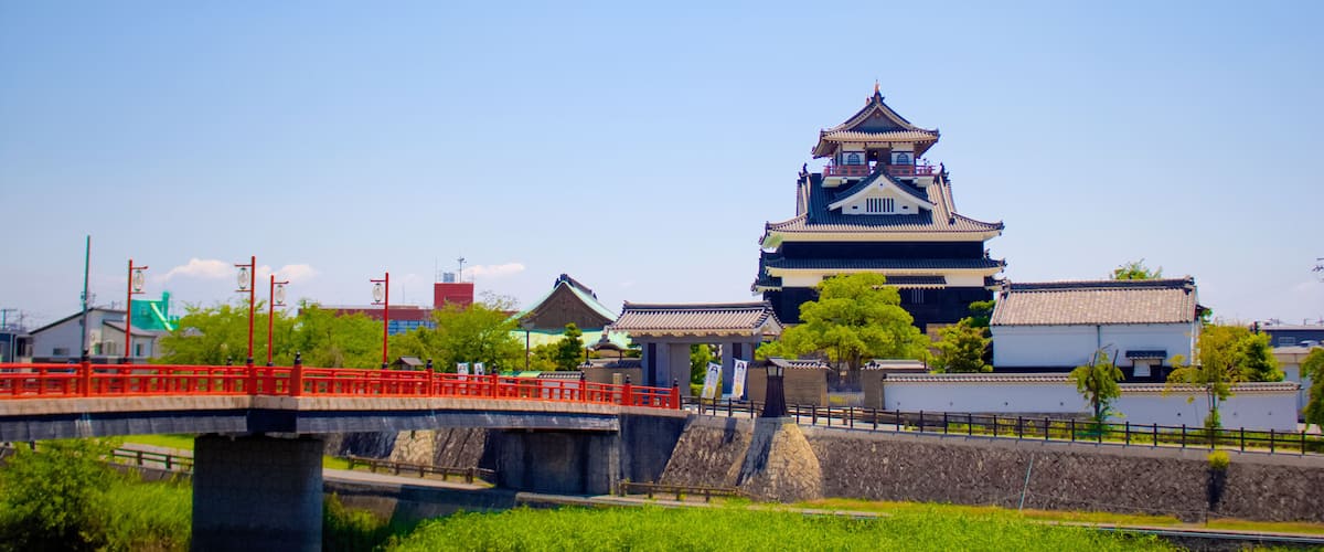 A traditional Japanese castle with a prominent tower, a stone bridge with red railings, and lush greenery.