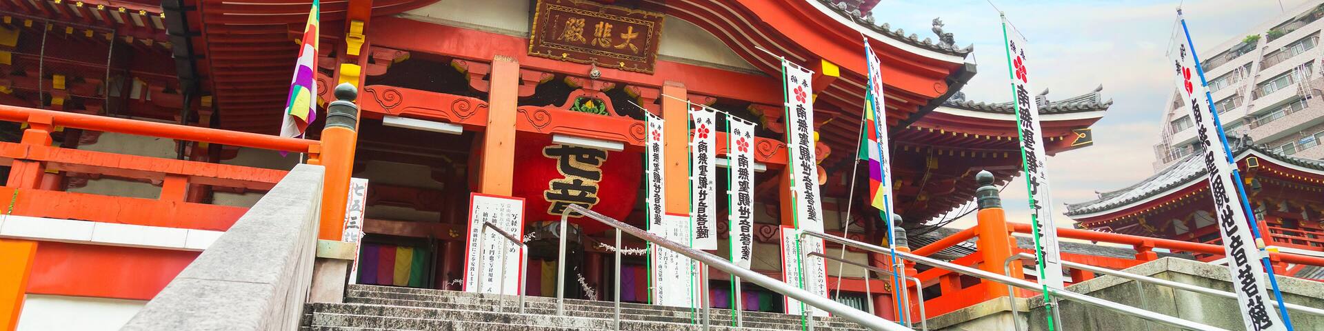 Un templo japonés tradicional con arquitectura roja y blanca, una escalera de piedra y banderas.