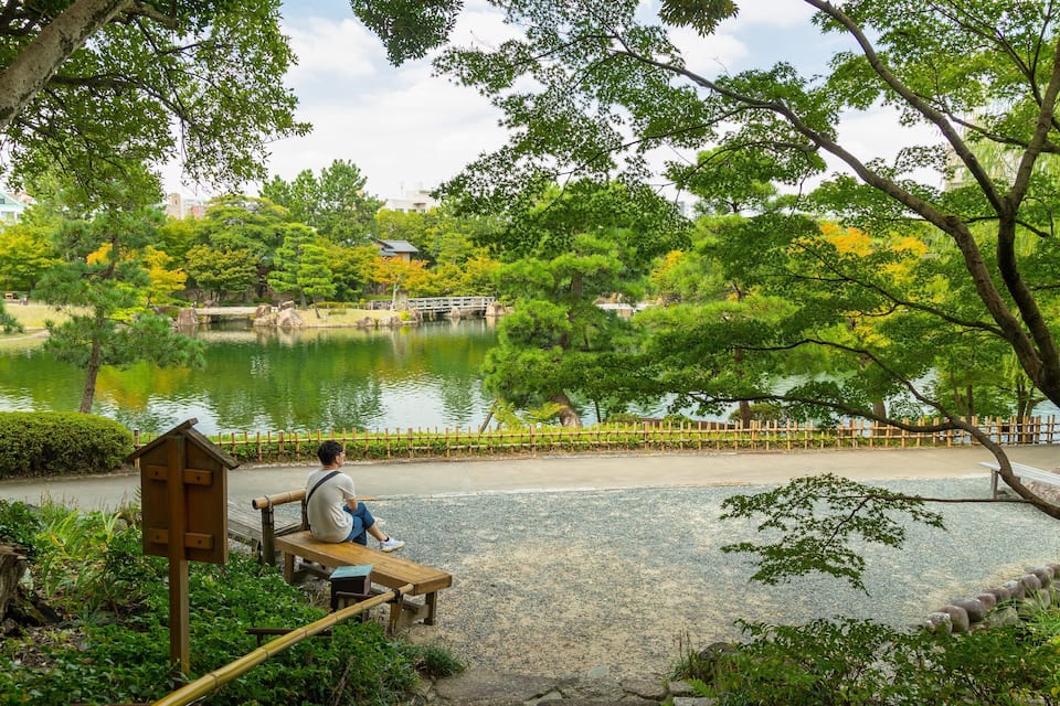 Ein friedlicher Park mit Teich, üppigem Grün, einer Holzbrücke und einer Person, die auf einer Bank sitzt.