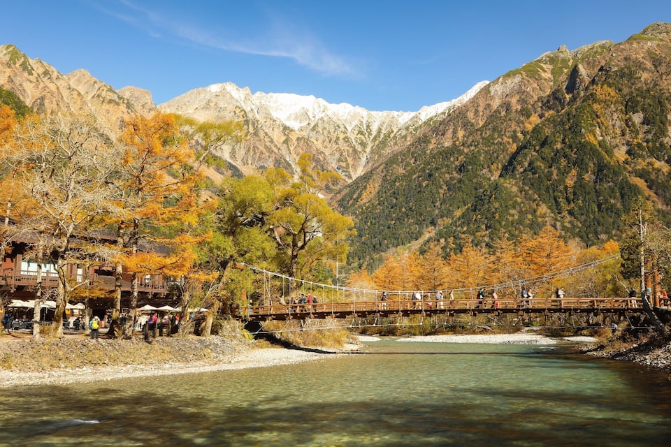 Un río con un puente de madera, árboles de colores otoñales y montañas al fondo.
