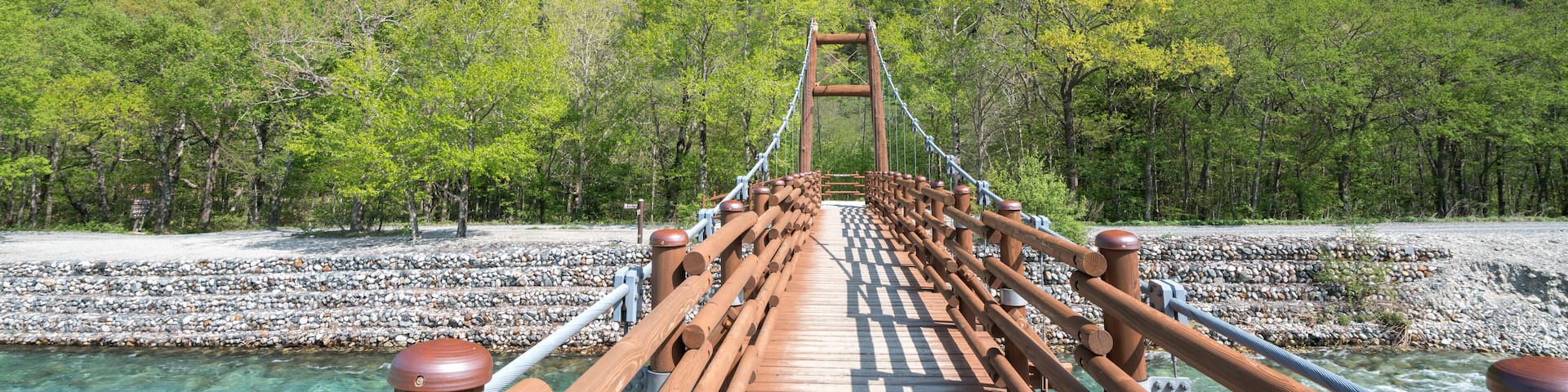 A wooden suspension bridge over a clear river with rocky banks, surrounded by dense green forest and mountains in the background.