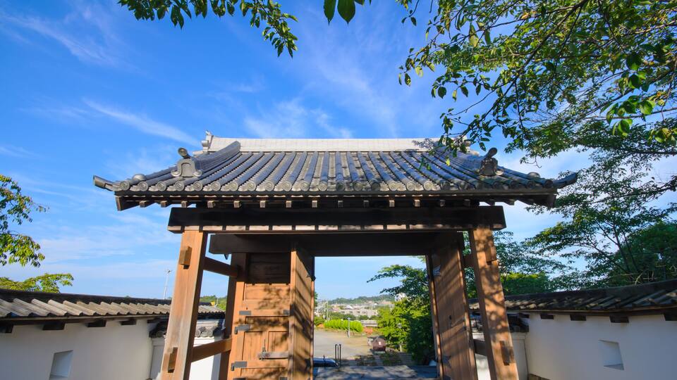 A traditional Japanese gate with a tiled roof, surrounded by white walls and a pathway leading through it.