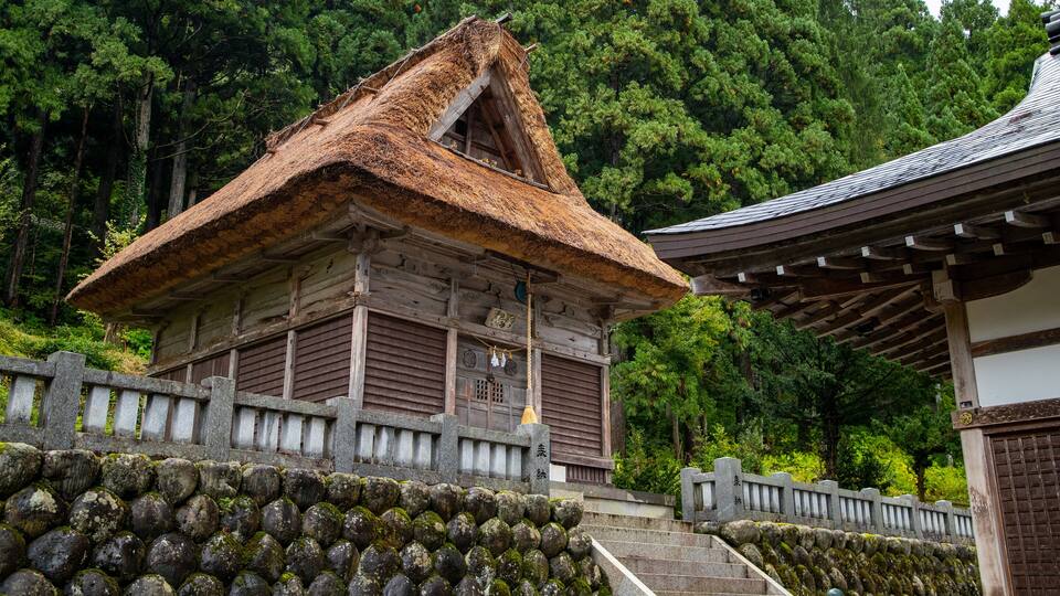A traditional Japanese building with a thatched roof, surrounded by a stone wall and lush greenery.