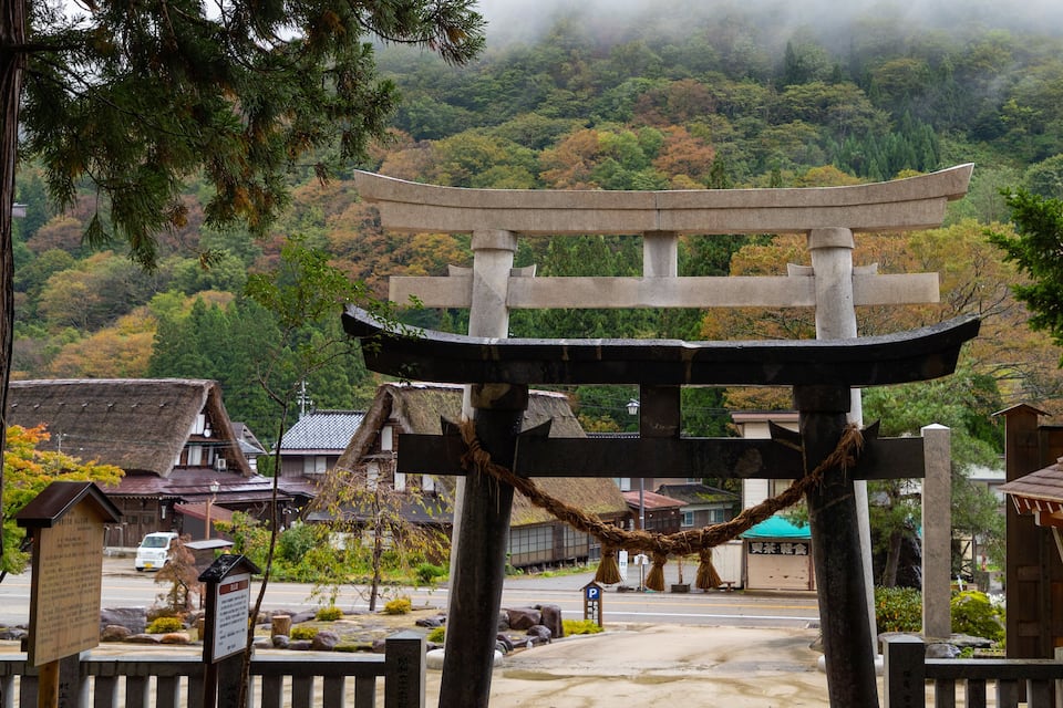 A traditional Japanese torii gate with a rope tied around it, surrounded by buildings and a forested hillside.