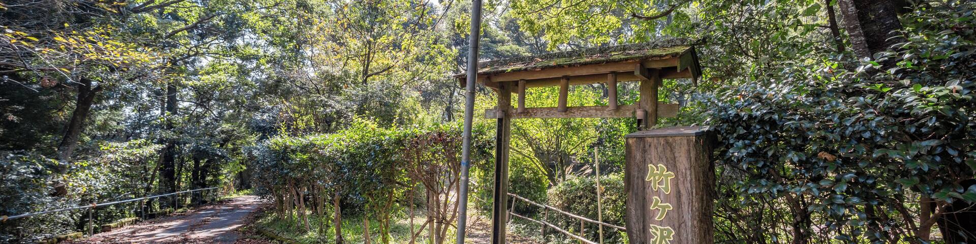 Zona boscosa con un camino pavimentado, un arco de madera y un letrero con caracteres chinos.