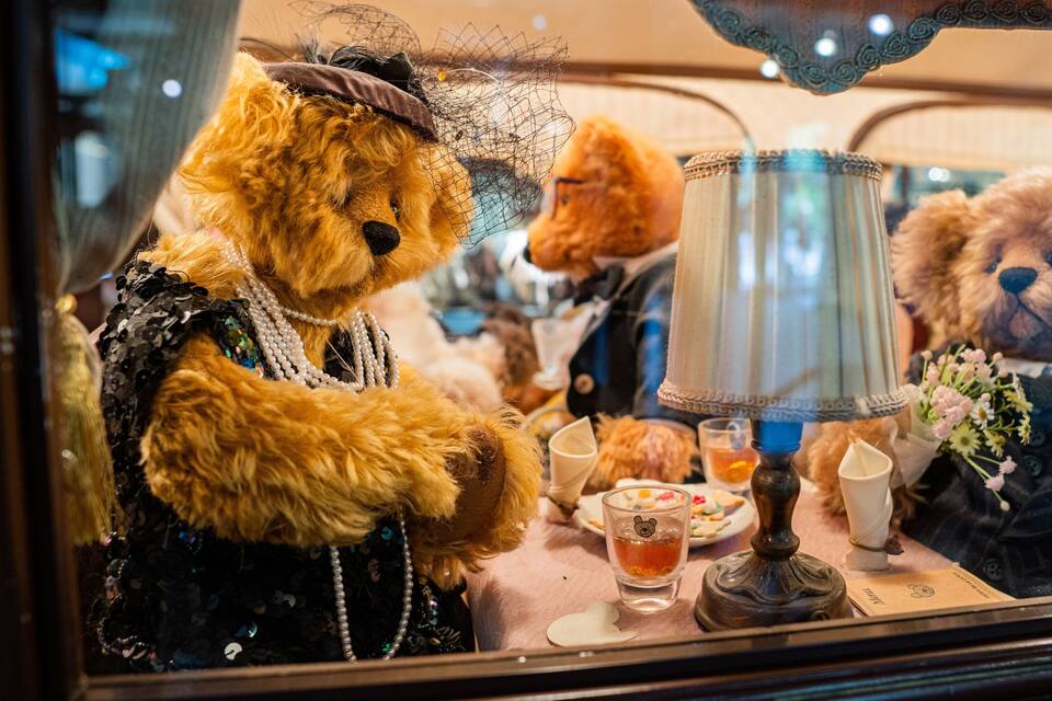 Un ours en peluche, portant un chapeau et une robe, est assis à une table avec une lampe et une tasse de thé.