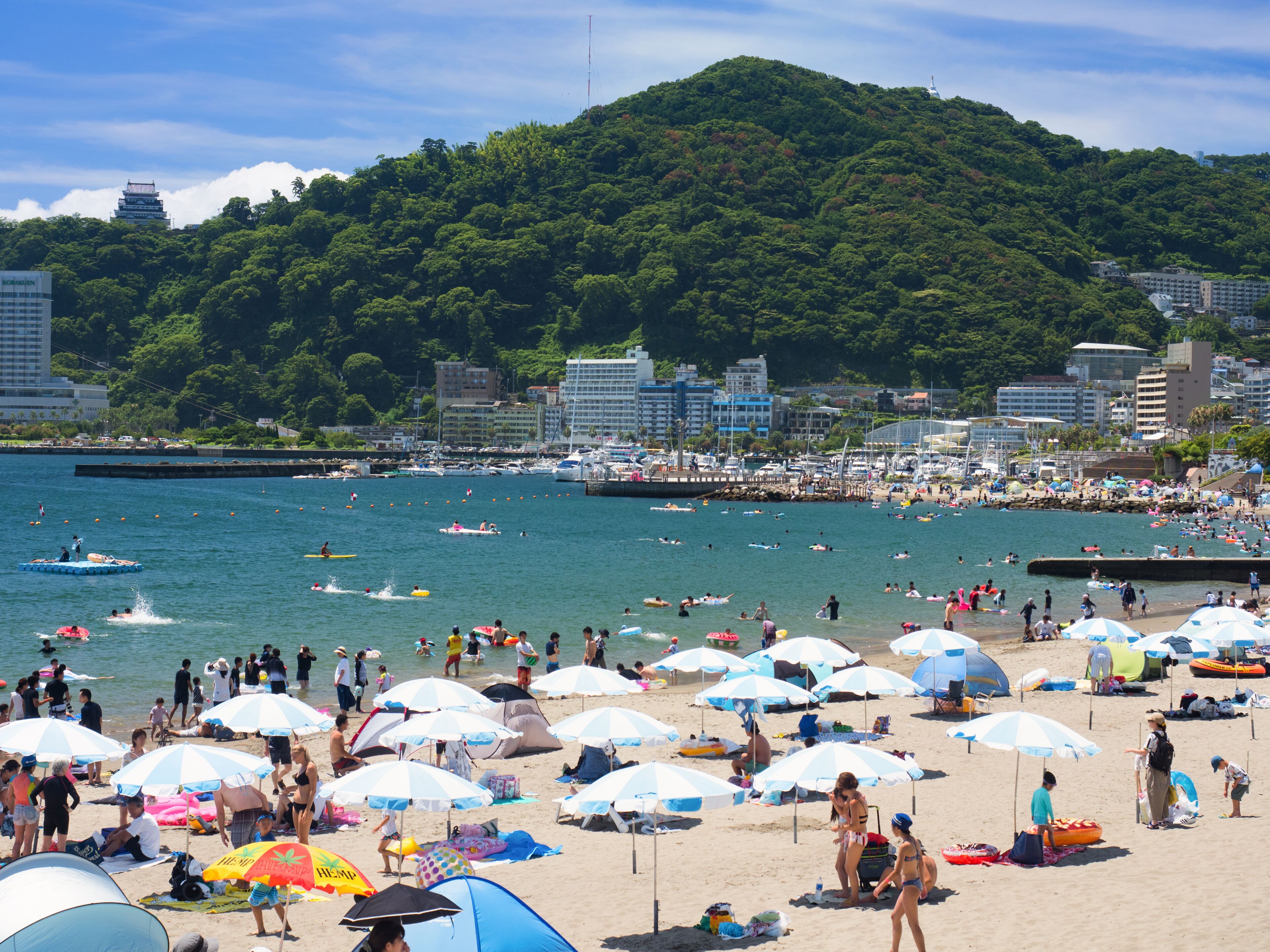 A crowded beach with numerous umbrellas, people sunbathing, and boats on the water.
