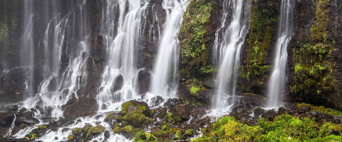A multi-tiered waterfall surrounded by lush greenery and rocky terrain.