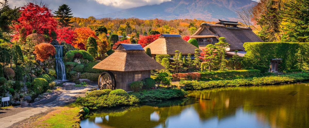A traditional Japanese garden with a waterfall, thatched-roof buildings, and a mountain in the background.