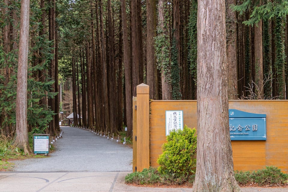 A forested pathway leading to a memorial park entrance with a wooden gate and a signboard.