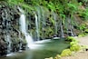 A multi-tiered waterfall cascading over moss-covered rocks into a calm river.
