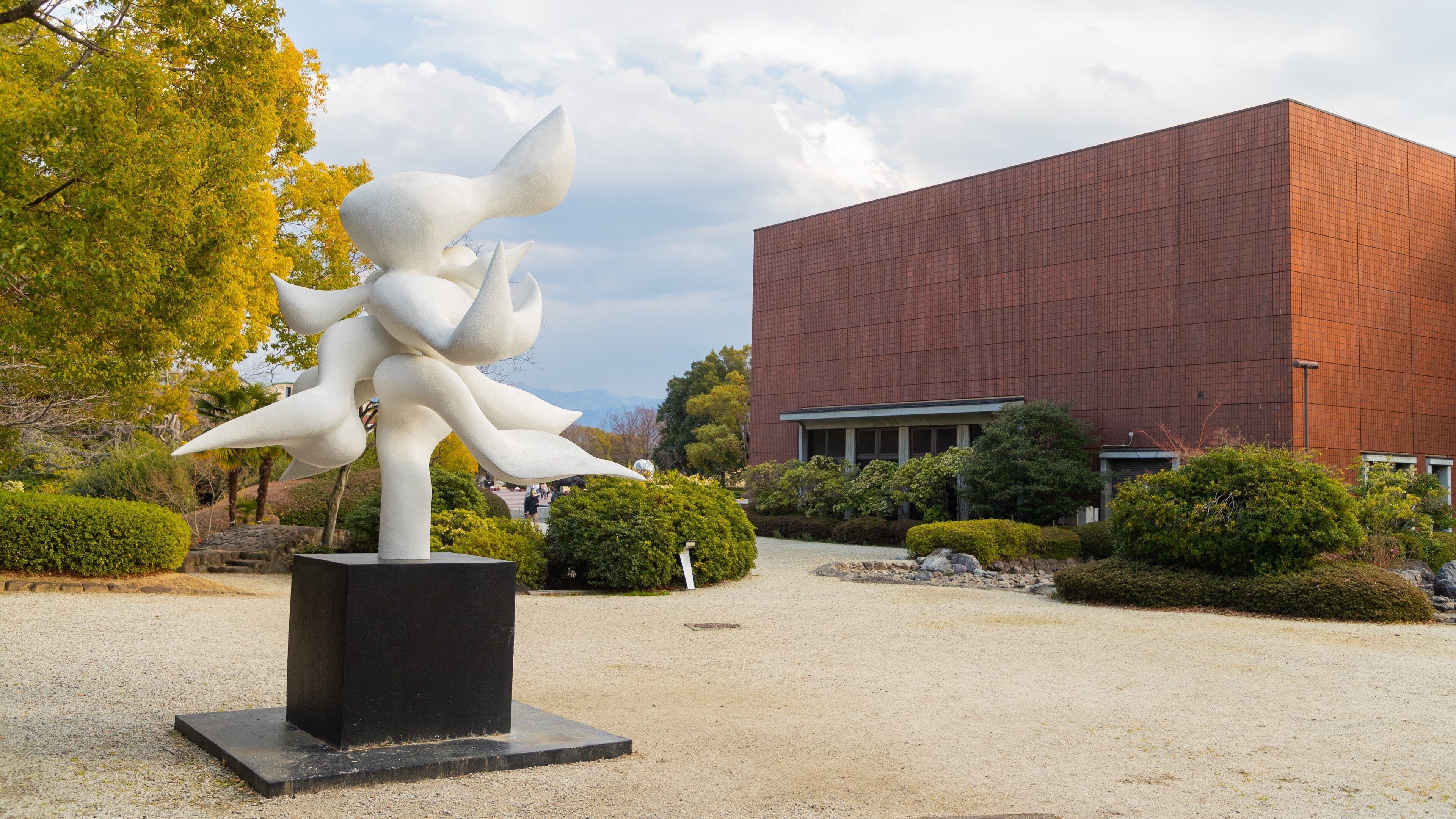 A sculpture of a white figure with multiple arms in a garden with a red building in the background.