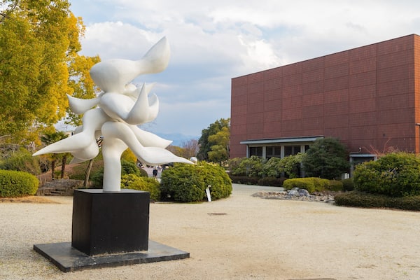 Une sculpture représentant une figure blanche dotée de plusieurs bras, dans un jardin, avec un bâtiment rouge en arrière-plan.