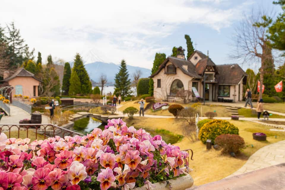 A garden with pink flowers, a bridge, and a house with a mountain in the background.