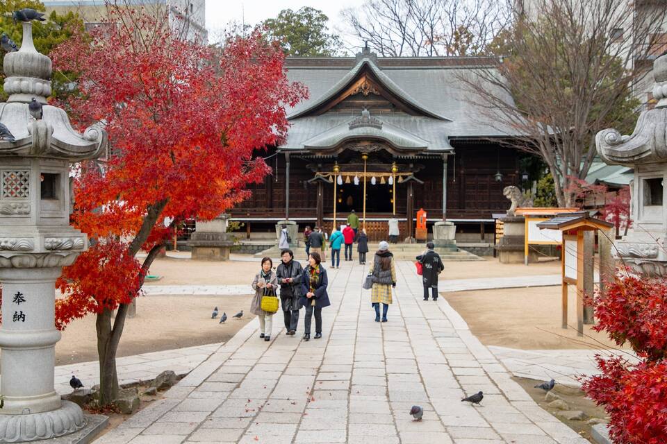 Et traditionelt japansk tempel med røde efterårsløv, stenenlanterner og folk, der går i gården.
