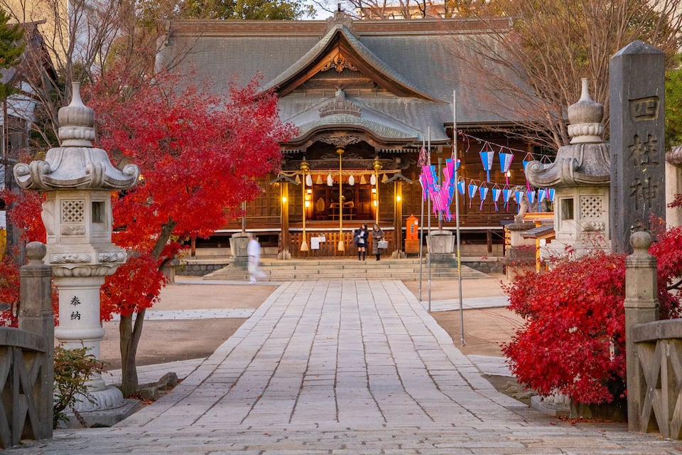 Um edifício tradicional japonês com entrada coberta por telhas, rodeado por folhagem avermelhada e lanternas de pedra.