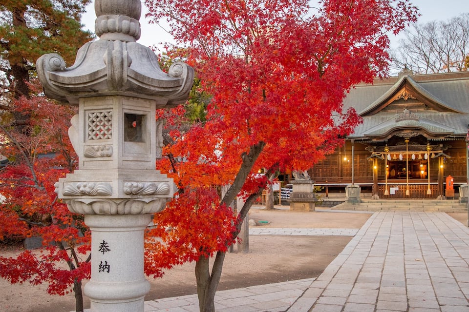 Um candeeiro de pedra com caracteres japoneses, folhas vermelhas de outono e um edifício tradicional ao fundo.