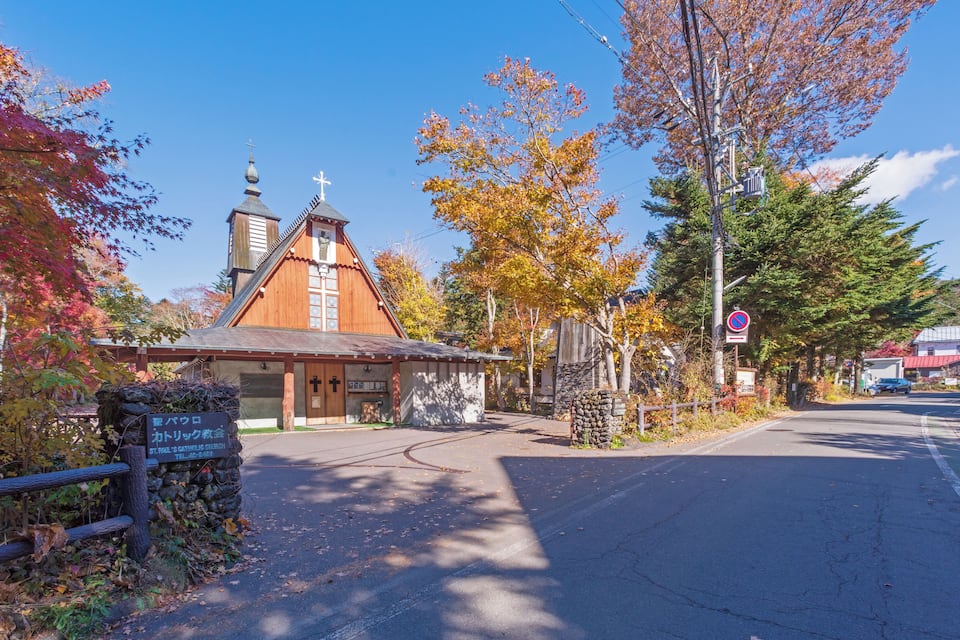 A church with a wooden facade and a cross on top, surrounded by autumn trees and a clear sky.