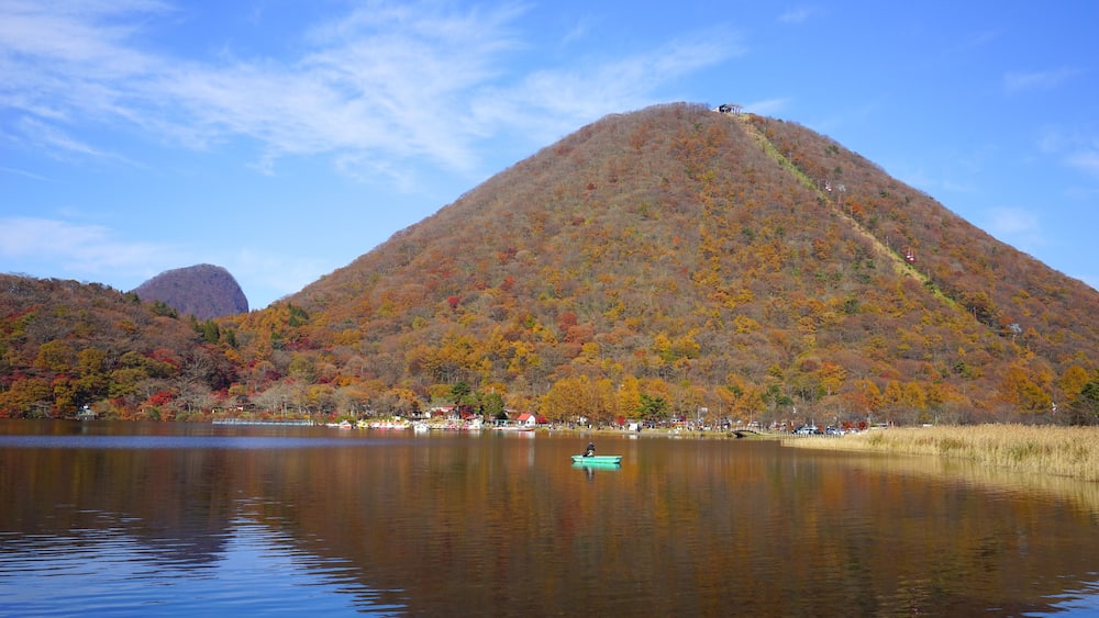 A calm lake with a green boat, a mountain with autumn foliage, and a clear blue sky.