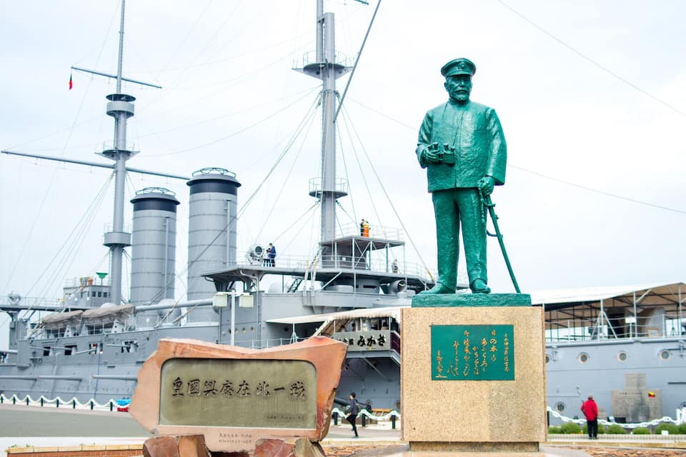 Une statue d’un marin se trouve devant un grand navire, et une pancarte est placée devant.