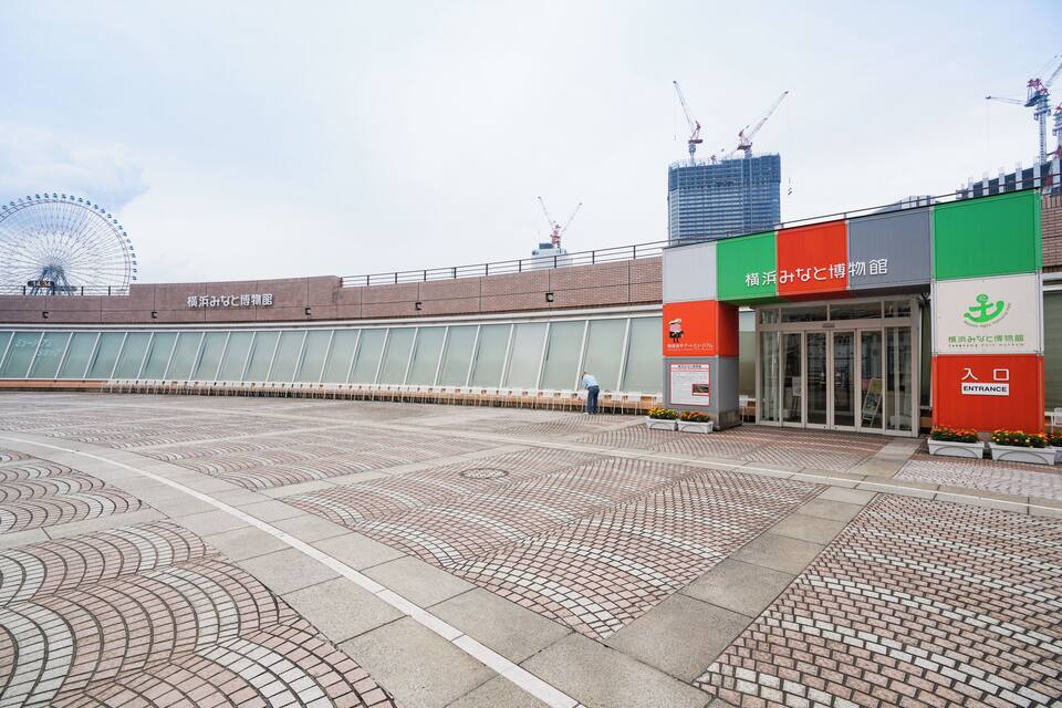 A spacious plaza with patterned paving, a building with a green and red sign, and a Ferris wheel in the background.
