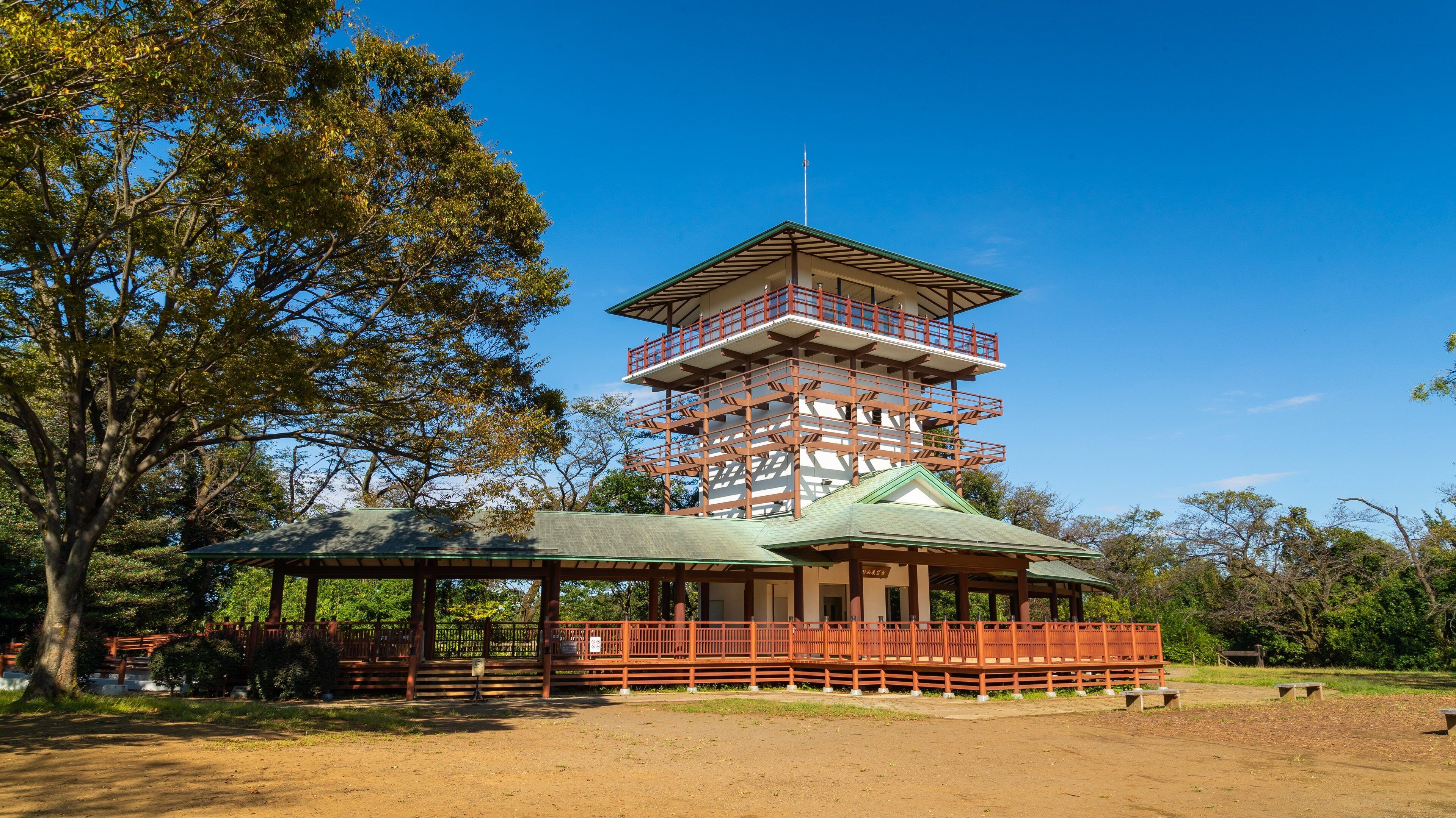 Una pagoda japonesa tradicional con techo verde y barandillas de madera, rodeada de árboles y un sendero de tierra.