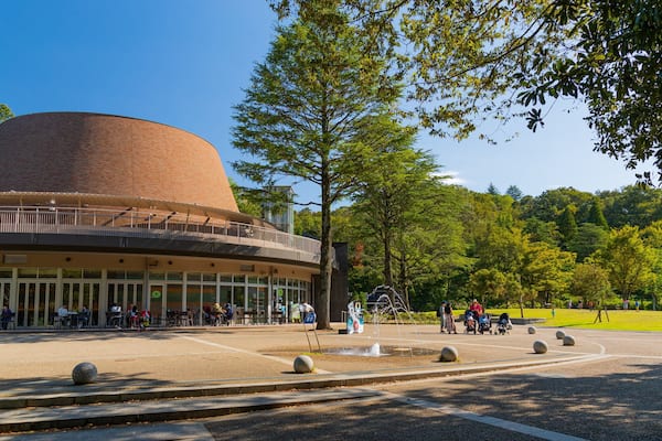 A modern building with a curved roof, surrounded by a park with trees and a fountain.