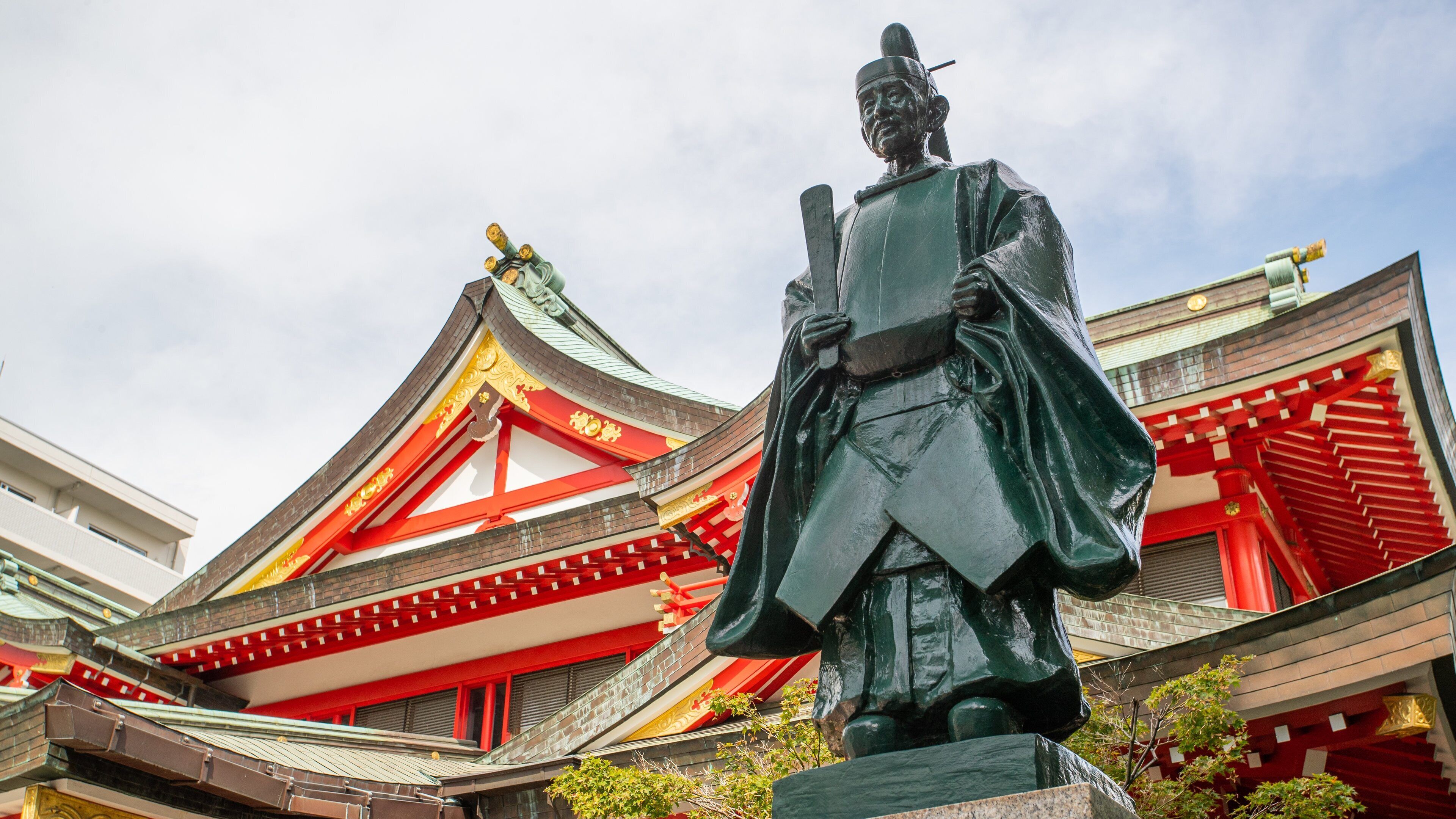 Una estatua de una figura histórica vestida con atuendo tradicional, frente a un edificio tradicional con detalles en rojo y dorado y un tejado verde.