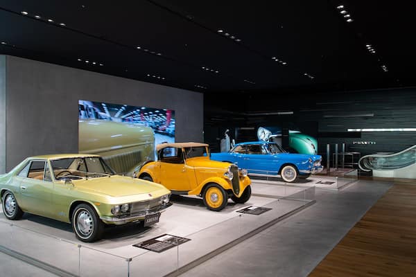 Vintage cars on display in a modern museum with a large digital screen in the background.