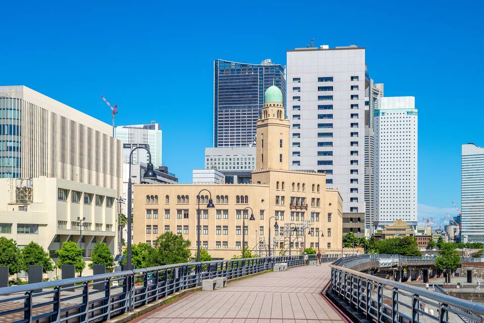 Un paysage urbain avec des bâtiments modernes, une tour imposante surmontée d’un dôme et un pont piétonnier.