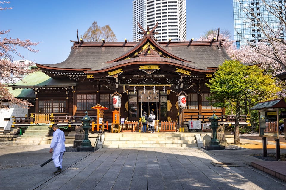 A traditional Japanese temple with a curved roof and wooden structures, surrounded by cherry blossoms and modern buildings in the background.