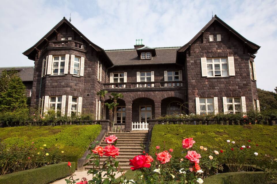 A large, dark-stone house with white shutters and a well-manicured garden with roses.