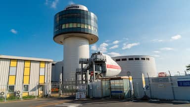 Un vaste complexe industriel comprenant une tour cylindrique imposante, un grand bâtiment blanc orné d’une bande rouge et une zone délimitée par une clôture, où se trouve un arrêt de bus.