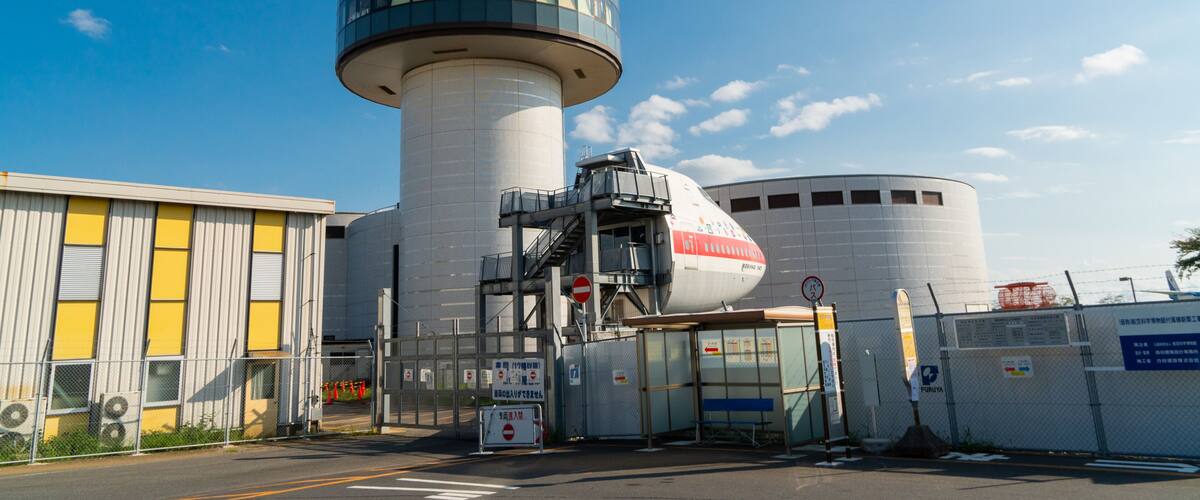 A large industrial facility with a prominent cylindrical tower, a large white building with a red stripe, and a fenced area with a bus stop.