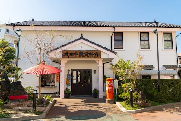 Un bâtiment traditionnel japonais, avec un toit recouvert de tuiles et un parapluie rouge devant l’entrée.