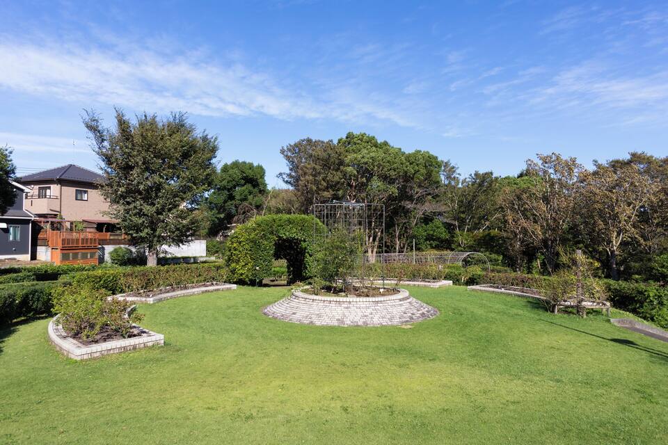 Un jardí ben cuidat amb una estructura circular de pedra, diversos arbres i una casa al fons.